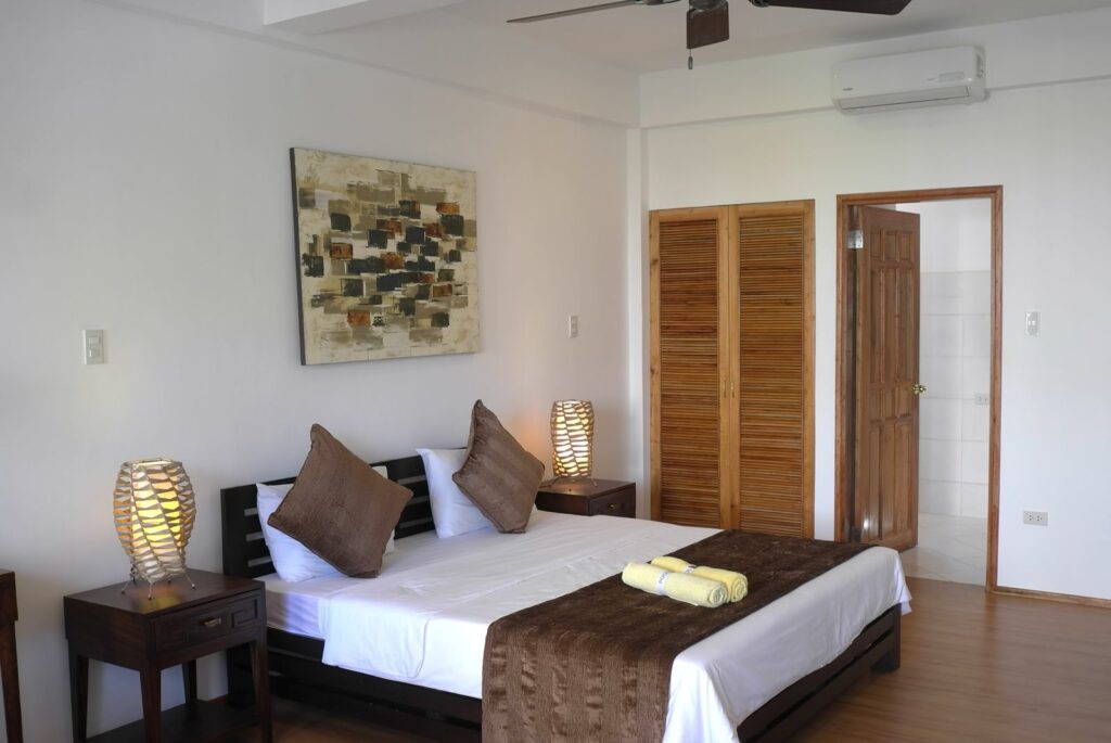 A neatly made bed with brown and white bedding, two large brown pillows, and two bedside tables with decorative lamps in a bright, modern bedroom. There's a wooden wardrobe, art on the wall, and an open door to a bathroom.