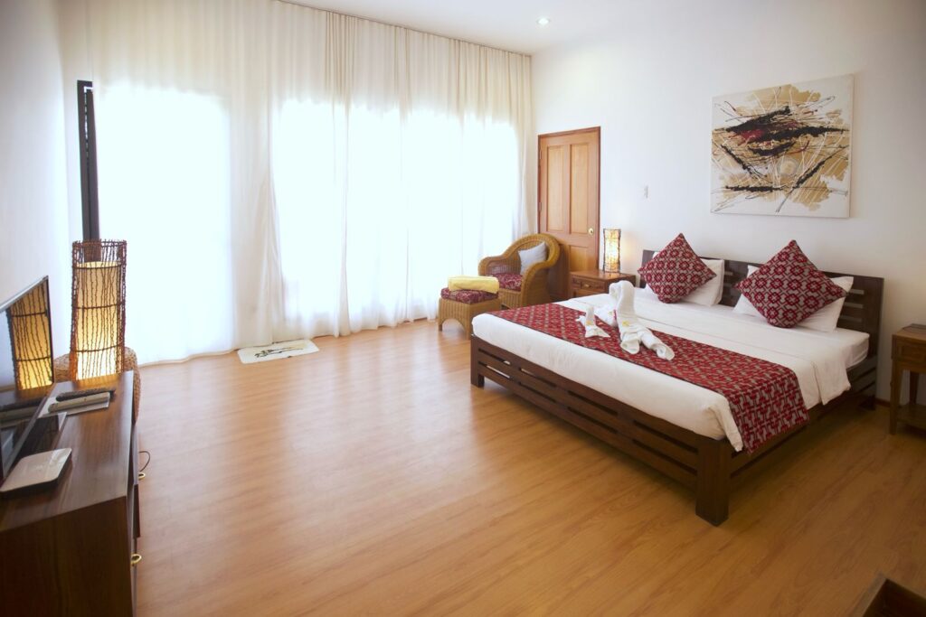 A spacious, bright hotel room with a wooden floor, a double bed with red and white pillows and blanket, a TV, an armchair by the window, and white curtains letting in natural light. Towels are folded on the bed.